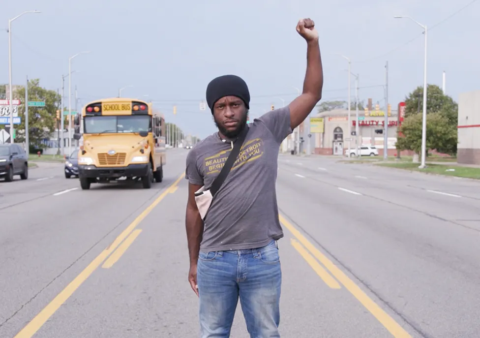 A Black man stands in the middle of a turn lane on a six lane highway with his left arm raised in a fist.