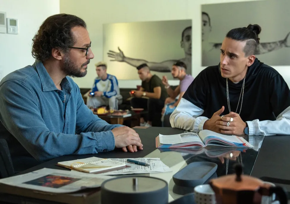 Two men sit at a table covered in books and papers and engage in a discussion.