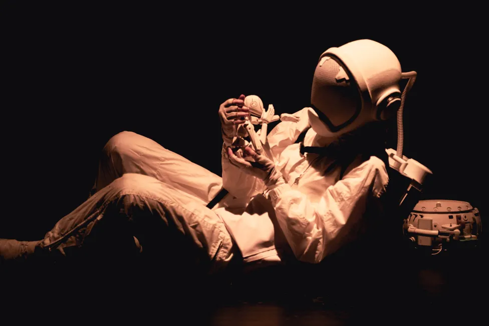 A person dressed as an astronaut lounges in a dark space while cradling a puppet in their hands.