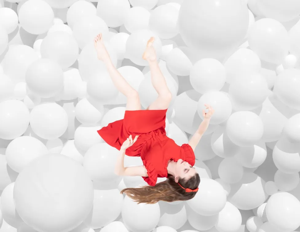 A woman in a red dress shown fallen down on a mass of white balls of multiple sizes.