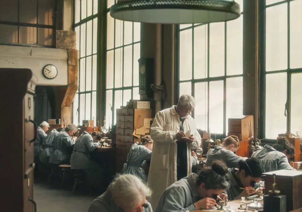 Employees in matching blue lab coats work together hunched over long tables.