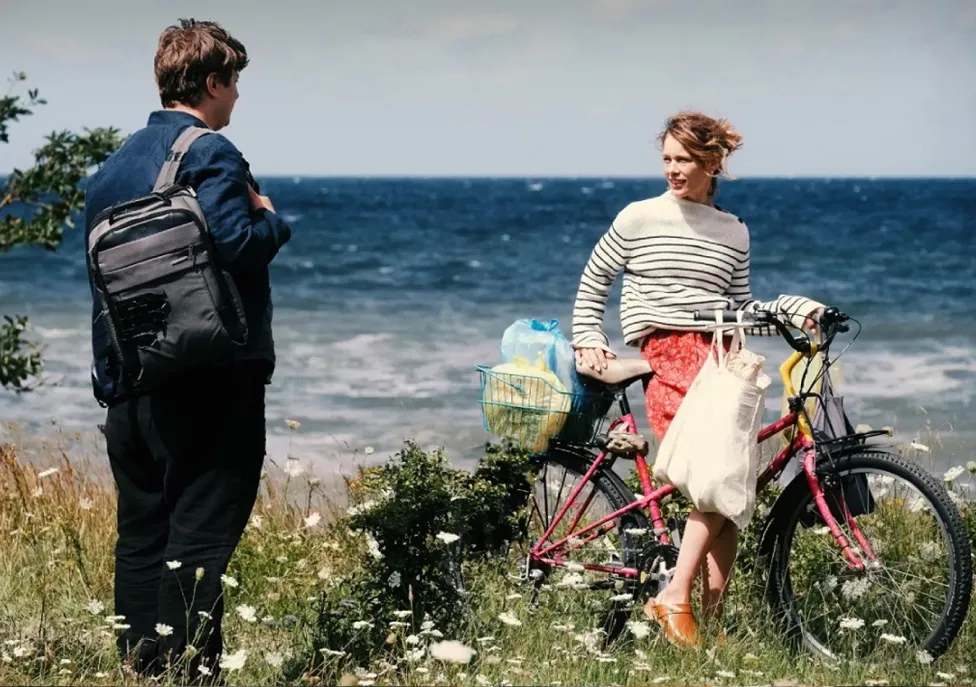 A woman sits on a bike in a flowery field in front of a beach, while speaking to a man with a backpack.