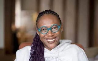Nandi Comer, smiling with braided hair and turquoise circle-framed glasses.