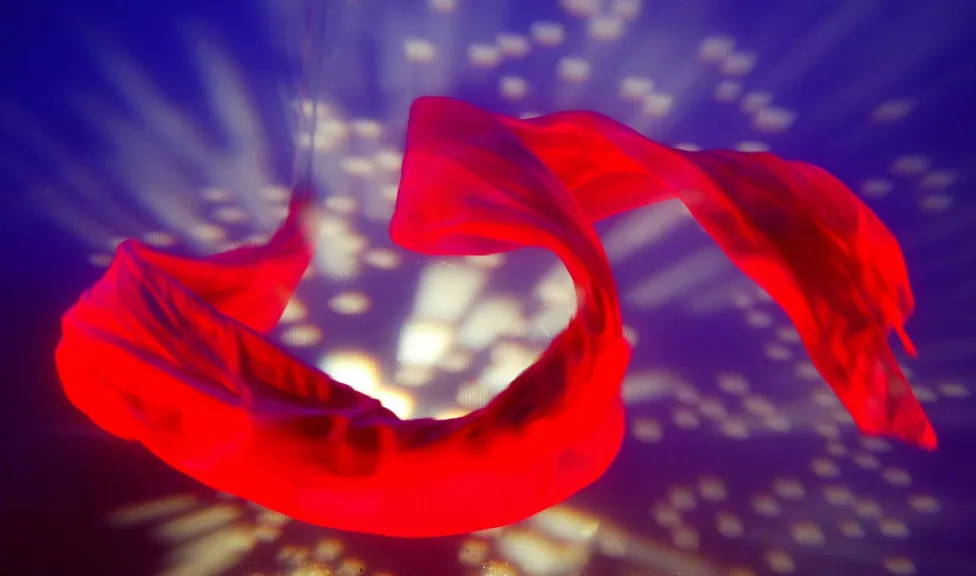 A red ribbon of silky fabric swirls through the air on a background of dark blue with gold sparks.