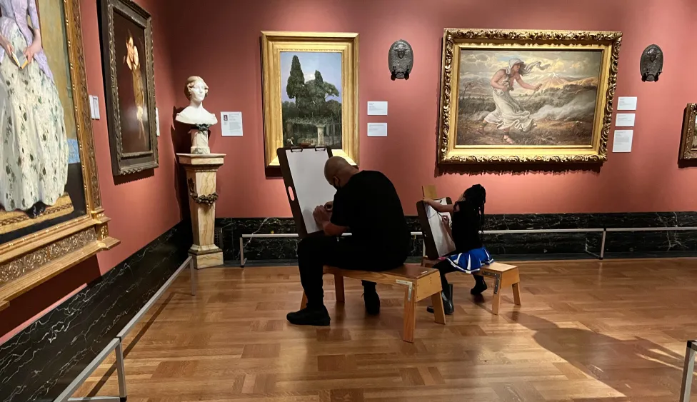 A man and young girl sit on easel benches drawing in the American galleries