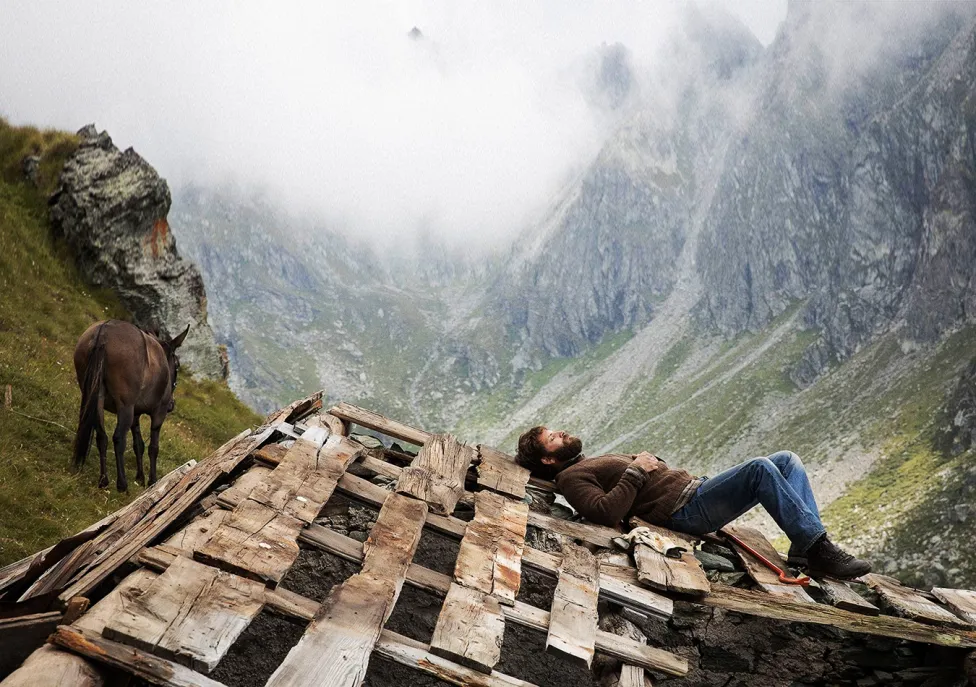 A man laying down on angled wood slats in the mountains with a horse nearby.