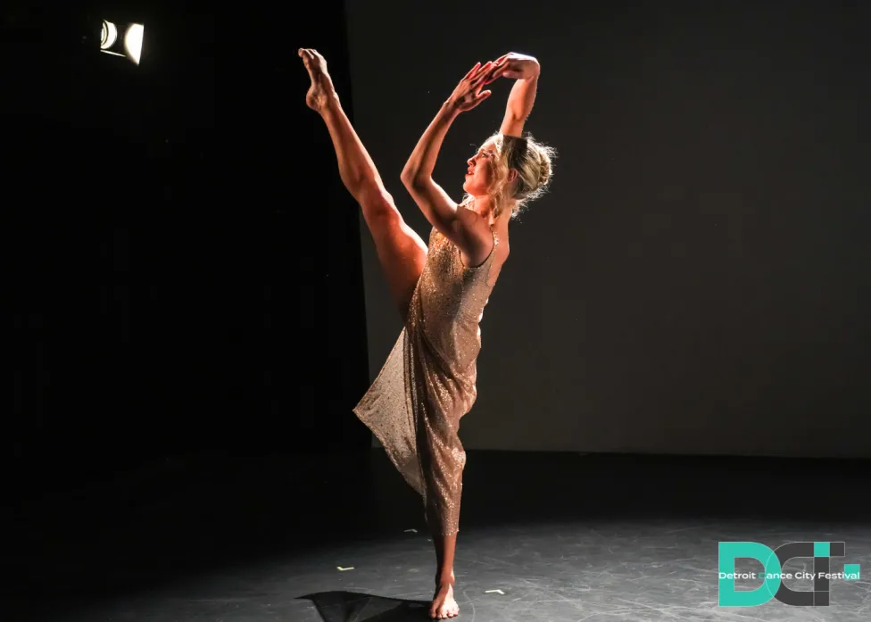 A lone dancer in a thin, beige dress holds her arms and one leg up in the air.