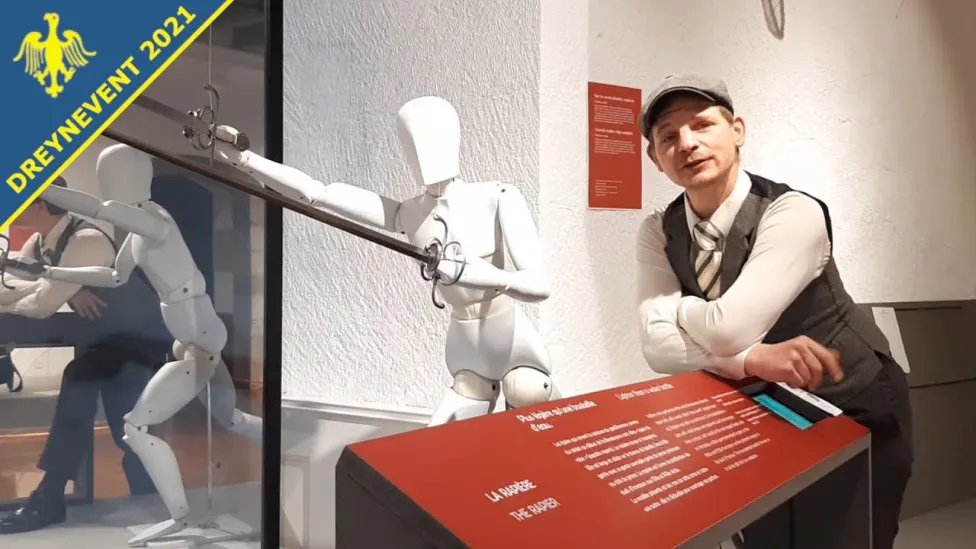 A man in a flat cap and vest poses in front of a museum sword display.