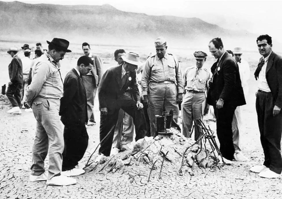 A group of men in suits and uniforms looking over a mound on the ground, in black and white.