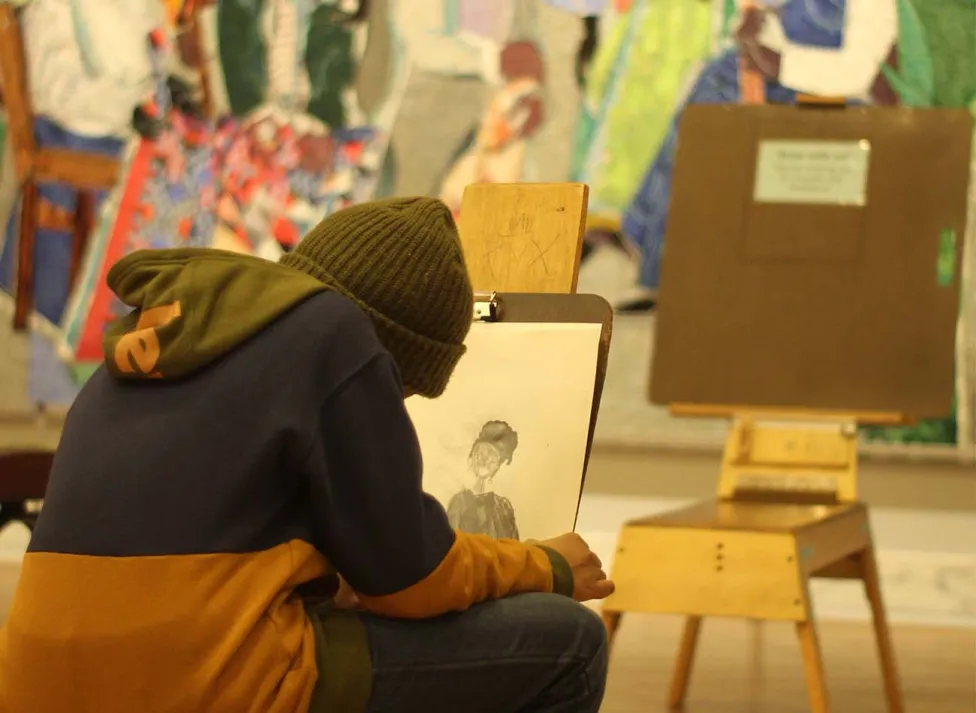 A person in a hoodie and beanie sits on an easel stool drawing in the *Contemporary galleries.