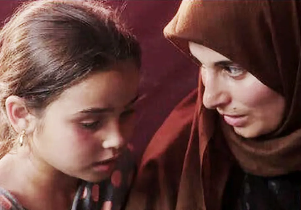 Two people keep their heads close together as they talk to one another. One is a younger girl and the other an older woman with a scarf around her head.