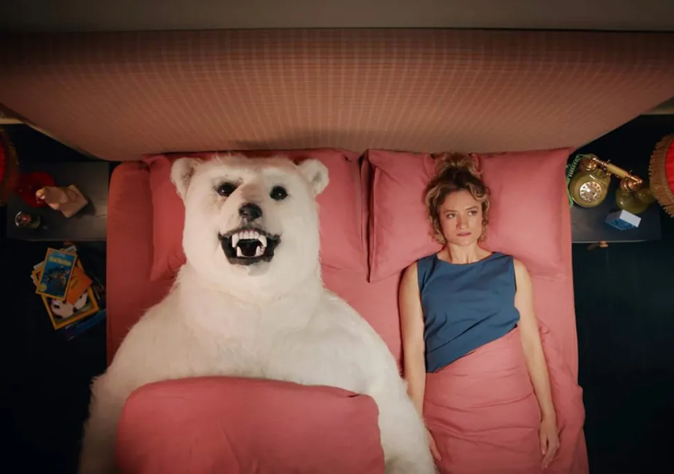 A woman and a polar bear lay on their backs on a bed, tucked into pink sheets.