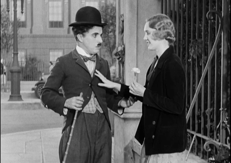 Charlie Chaplin stands next to a woman who's hand is on his chest, in black and white.