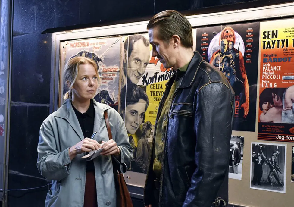 A man and a woman standing outside of a movie theatre bulletin board featuring film advertisements. 