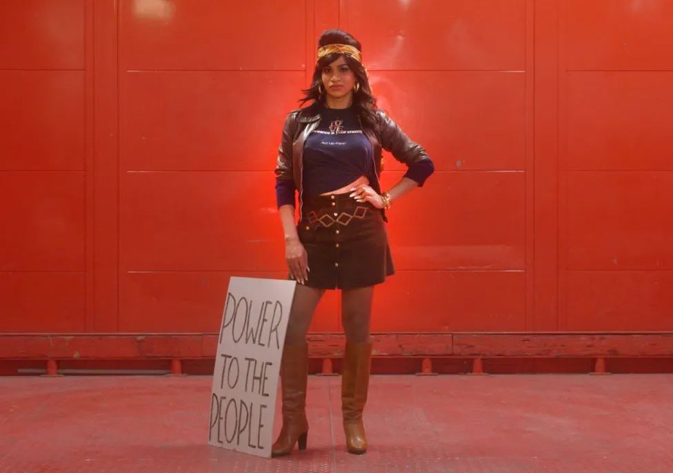 A woman in 70's style clothing and headband stands with a sign reading "Power to the people" in front of a red wall.