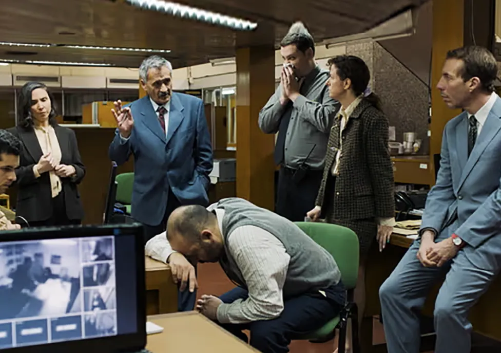 Five men and one woman wearing business suits gather around an office with shocked expressions.