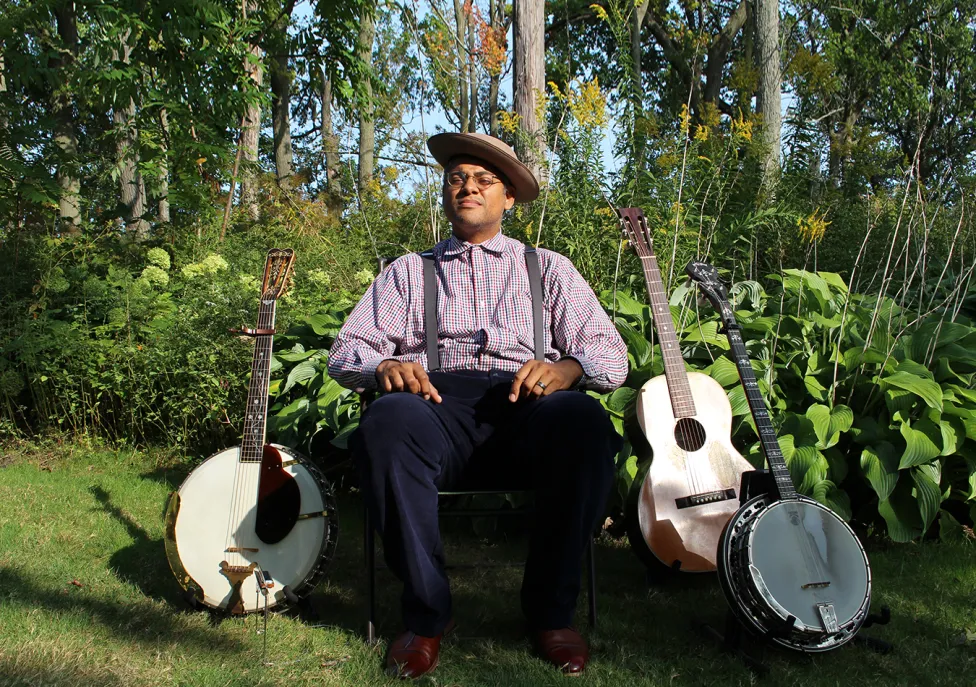 Don Flemmons sitting amongst greenery, surrounded by two banjoes and a guitar.