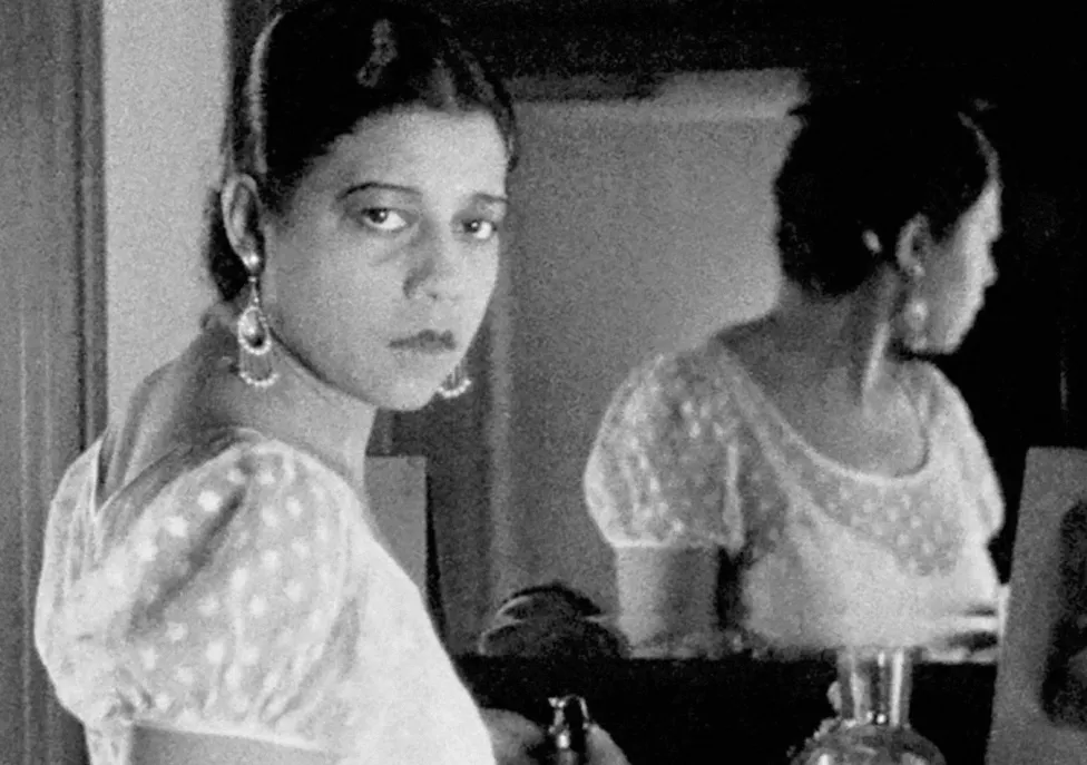 A woman stands in front of a mirror but looks back behind her towards the camera.