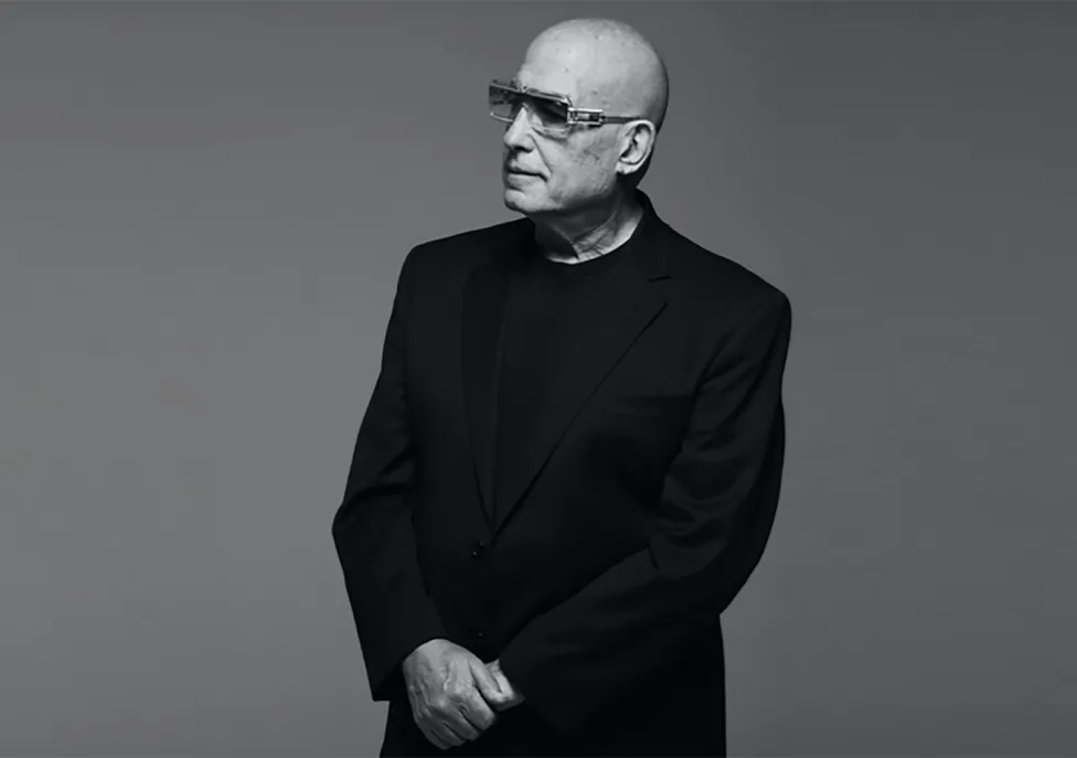 A bald man stands in a black suit wearing sunglasses.