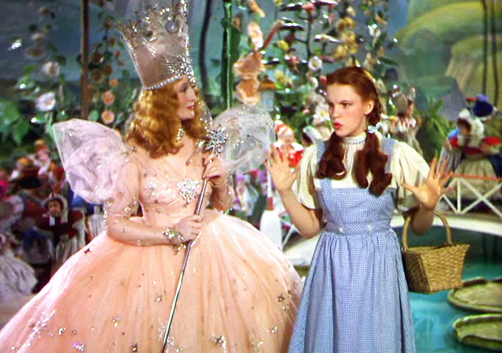 Scene from wizard of Oz, Glenda the good witch in a pink gown with wings next to Dorothy in a blue and white dress and pigtails.