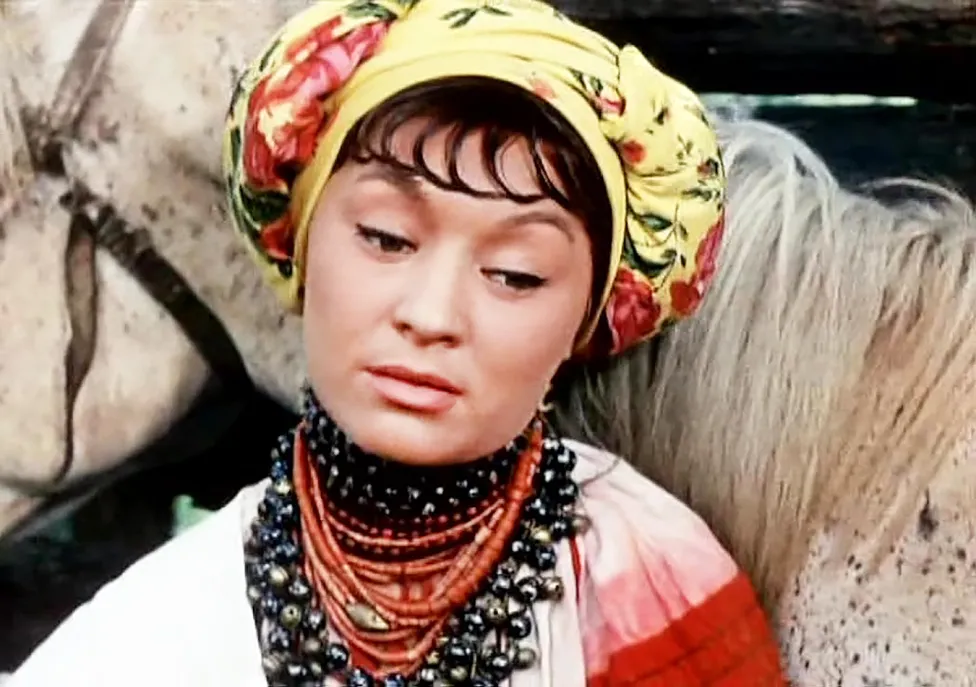 A woman in a yellow headscarf and chunky beaded necklaces stands in front of a white horse