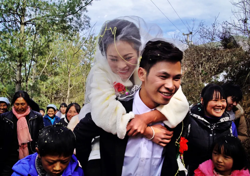 A woman in a bridal veil and white dress being held on the back of a man in a suit