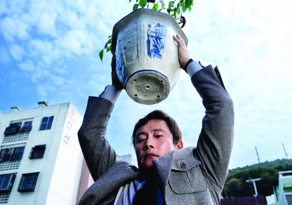 A man in a suit holds a potted plant high over his head while looking downward