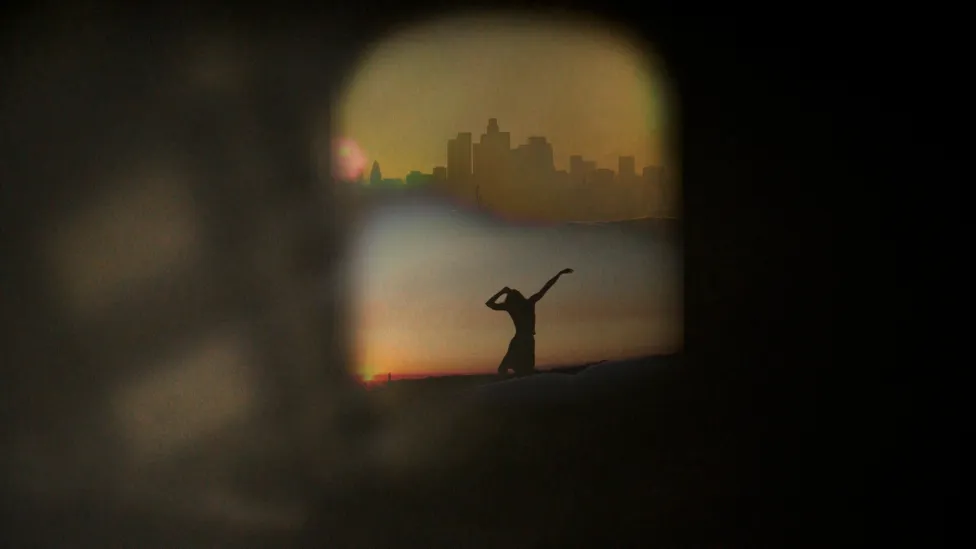Silhouette of single dancer in front of a city-scape, seen through a window surrounded by darkness