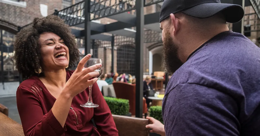A woman holds a glass of white wine as she laughs while chatting to a man in a backwards baseball cap.