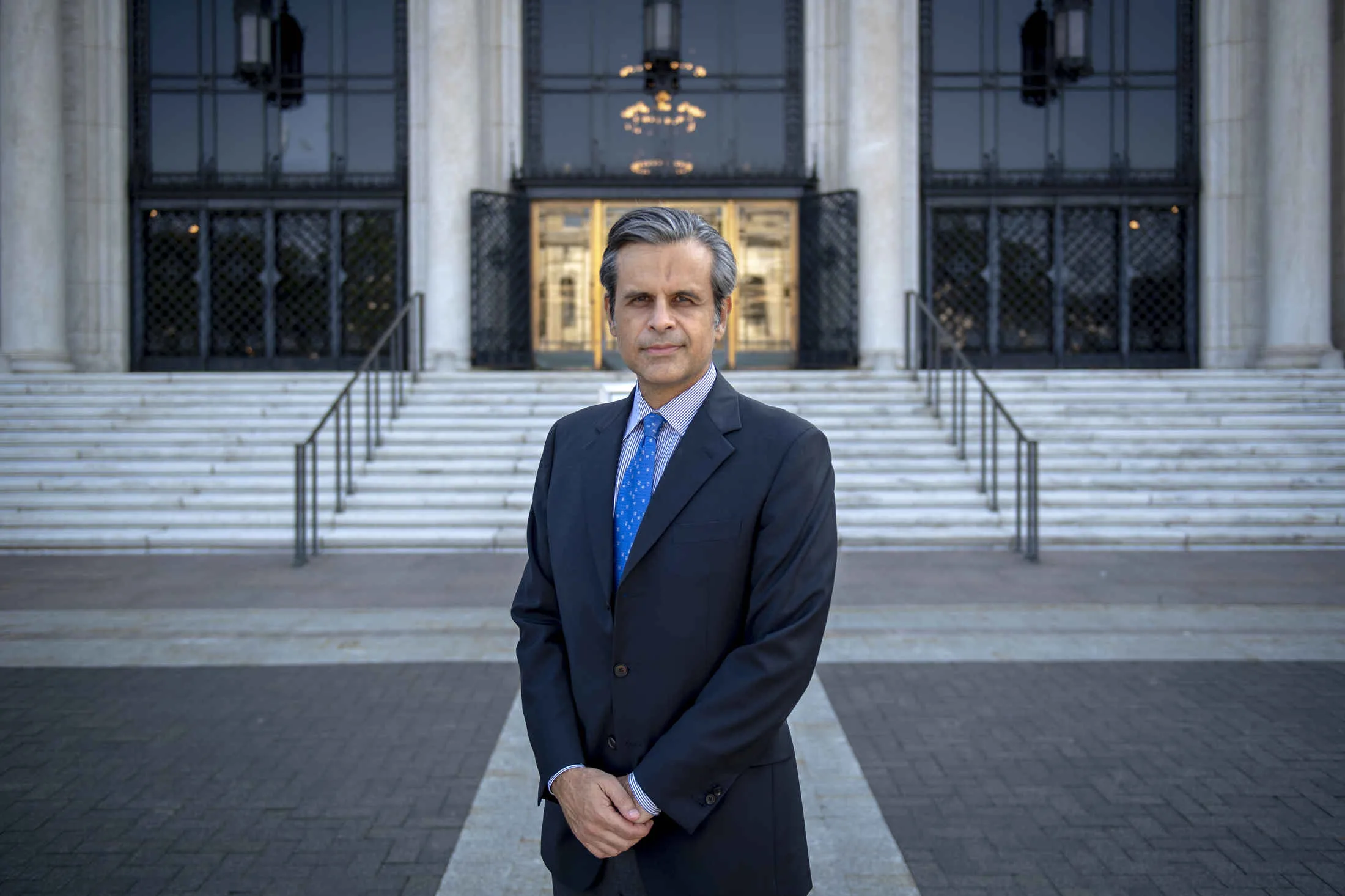 Salvador Salort Pons in a blue suit and tie, stands with his hands folded in front of himself in front of the front steps of the Detroit Institute of Arts