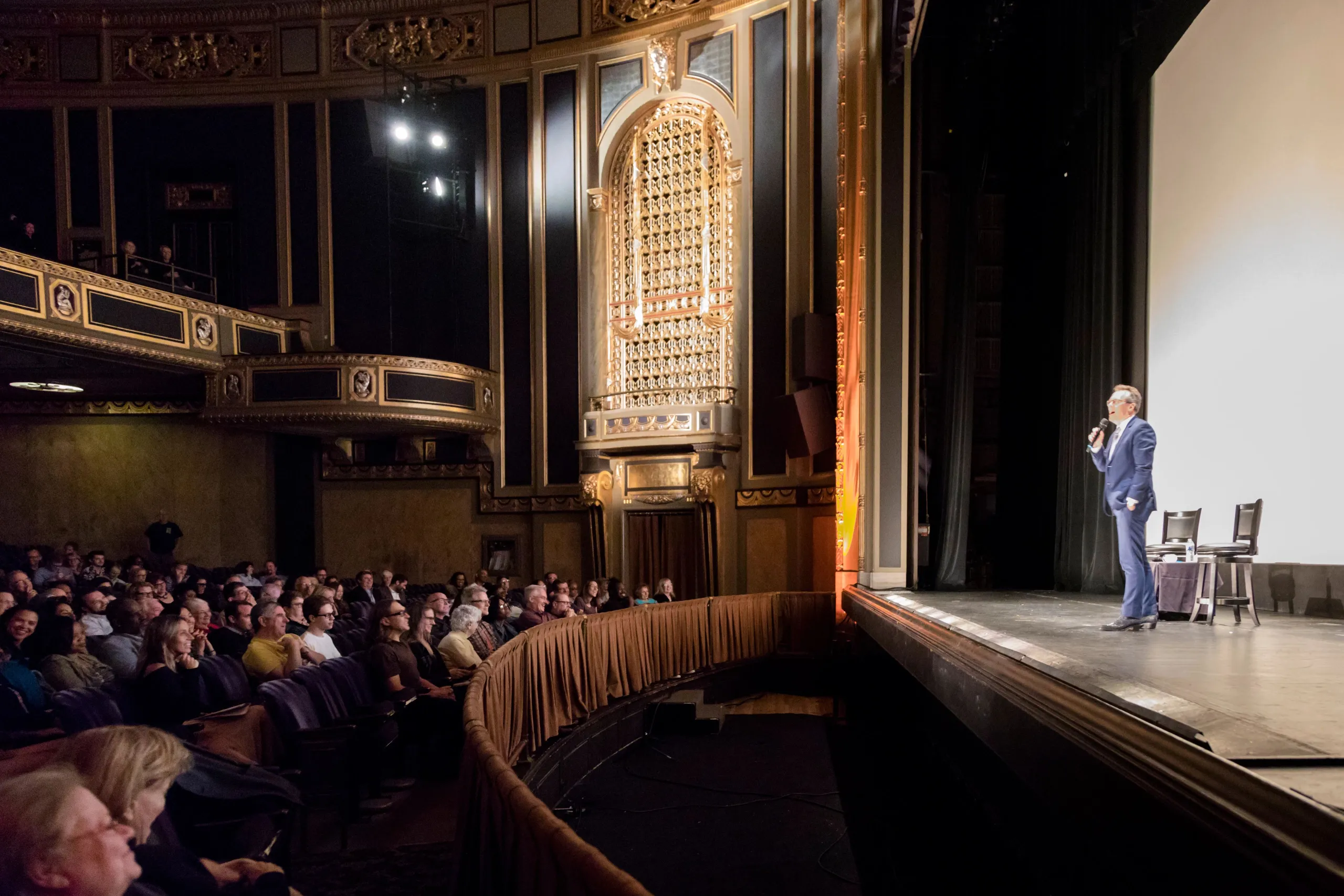Man in a suit speaking on stage at the Detroit Film Theatre auditorium