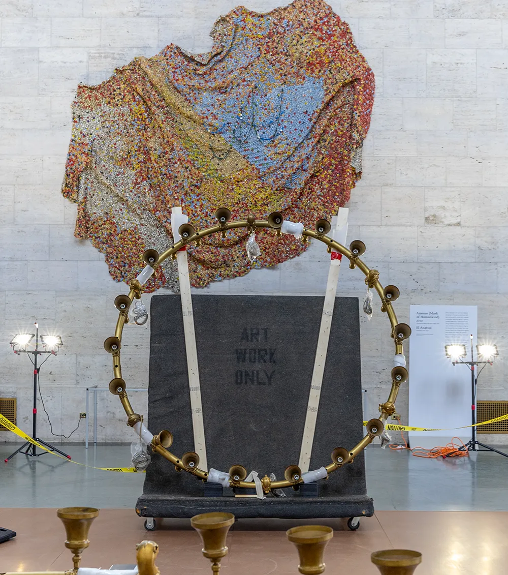 A chandelier secured on a large cart, aptly named “The Cadillac,” awaits transport, with Amemo (Mask of Humankind) by Ghanaian artist El Anatsui in the background, a generous loan from the collection of Padma and Raj Vattikuti.