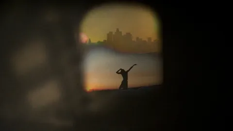 Silhouette of single dancer in front of a city-scape, seen through a window surrounded by darkness