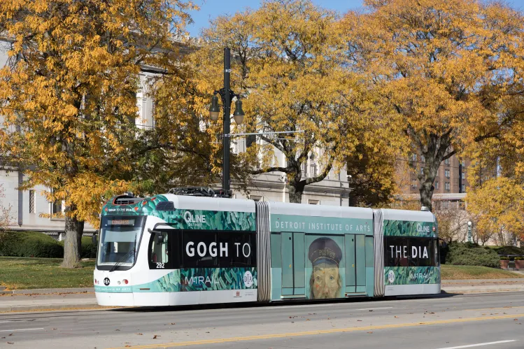 The Detroit QLine pictured amongst fall trees with a wrap featuring the DIA's iconic "Postman" by Van Gogh and the words Gogh to the DIA