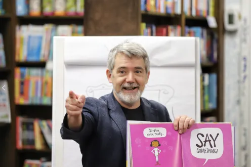 Author Peter Reynolds holding a book open to a bright pink spread and pointing to a crowd