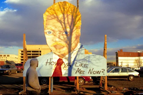 A person in a gray hoodie with the hood up stands shadowed in front of a large cutout sign of a head.