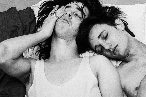 Two men lay next to each other with closed eyes and sleepy expressions, one shirtless, and the other in a white tank top.