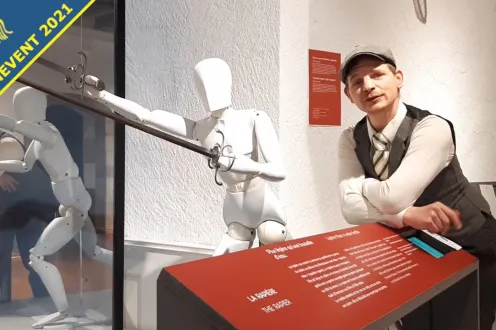 A man in a flat cap and vest poses in front of a museum sword display.