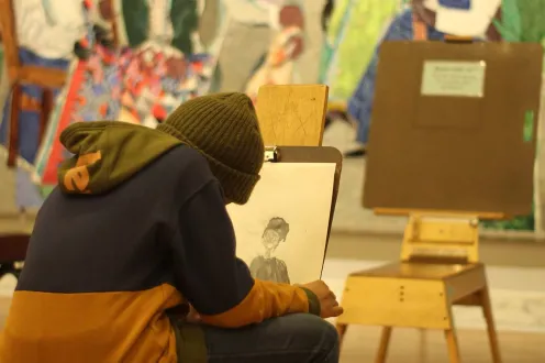 A person in a hoodie and beanie sits on an easel stool drawing in the *Contemporary galleries.
