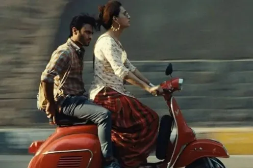 Two people, one in a dress, driving, and the other in jeans, sitting in the back, drive a red moped quickly down a street.