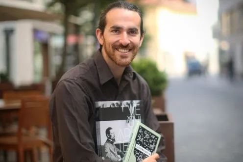 Noah Charney, a man with a short beard, sits wearing a large button down shirt and holding copies of his books in his arms.