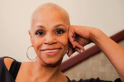 A woman with short cropped hair smiles at the camera with her head resting on her hand.