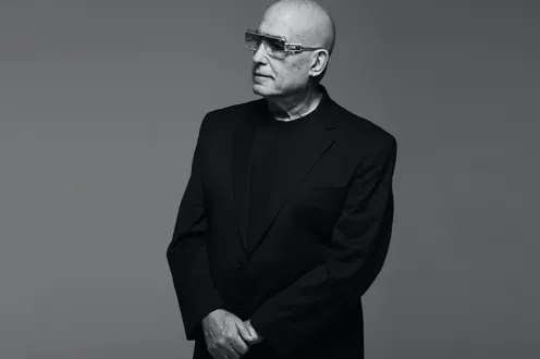 A bald man stands in a black suit wearing sunglasses.