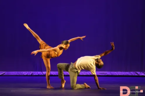 Two dancers on stage, one leaning towards the other and the other leaning away