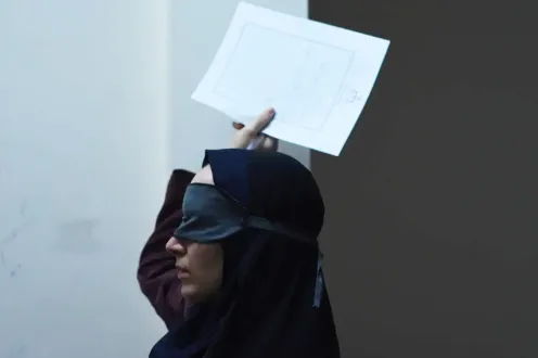In this still image from the film, Seed of the Sacred Fig, a woman wearing a black, silk blindfold holds a blank sheet of paper in the air.