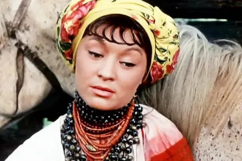 A woman in a yellow headscarf and chunky beaded necklaces stands in front of a white horse