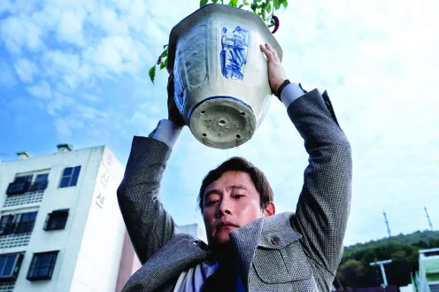 A man in a suit holds a potted plant high over his head while looking downward