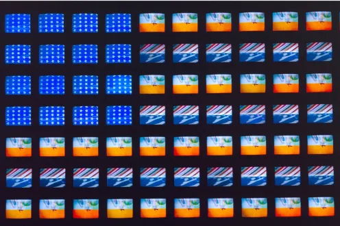 Image of Video Flag XGrid of small video screens arranged like a flag, displaying shifting patterns of blue fields with white stars and horizontal bands of red, white, and abstract imagery. The screens vary slightly in color and motion, creating a flickering, electronic interpretation of the American flag in Video Flag X by Nam June Paik.