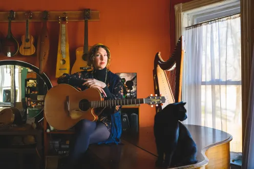 Audra Kubat seated on a piano holding an acoustic guitar with a black cat sitting in front looking out a window.