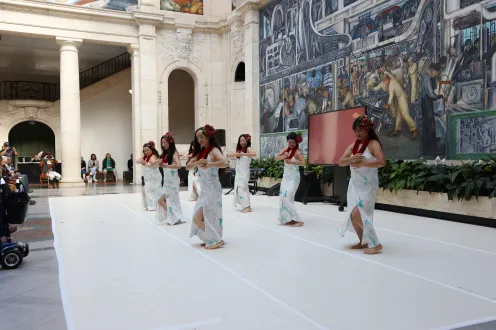 Group of hula dancers in long white dresses wearing maroon lays and flowers in hair.